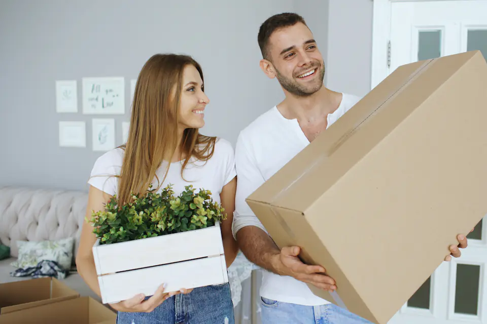 Couple carrying a large moving box and a potted plant into a bright hallway