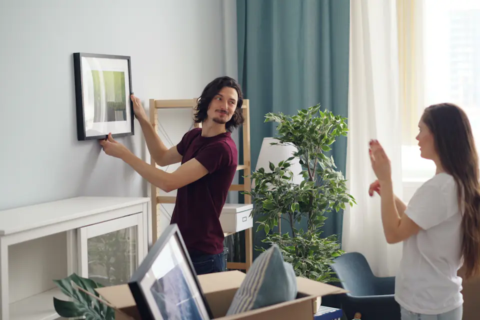 Man hanging a framed picture while a woman checks it is straight, with moving boxes nearby