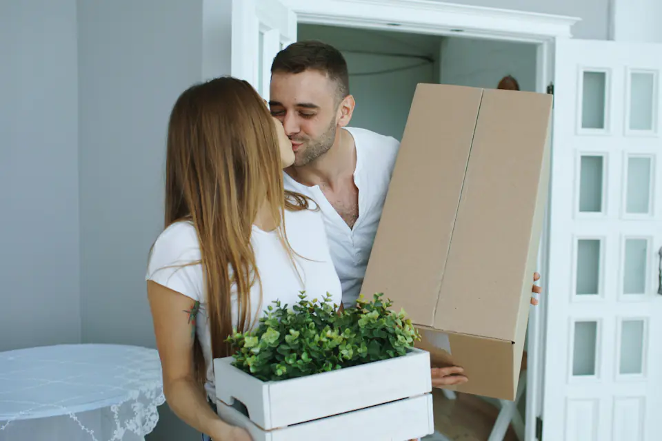 House removals in Newcastle Smiling couple carrying a large cardboard box and a planter through the door of their new home