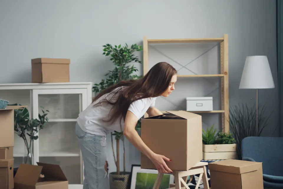 Woman lifting a moving box in a bright living room surrounded by packed boxes